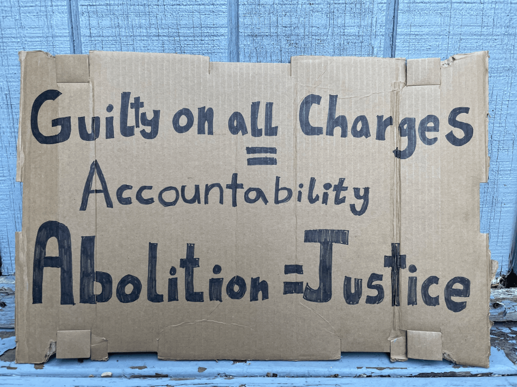 A cardboard sign made by Molly that says "guilt on all charges = accountability. Abolition = Justice."