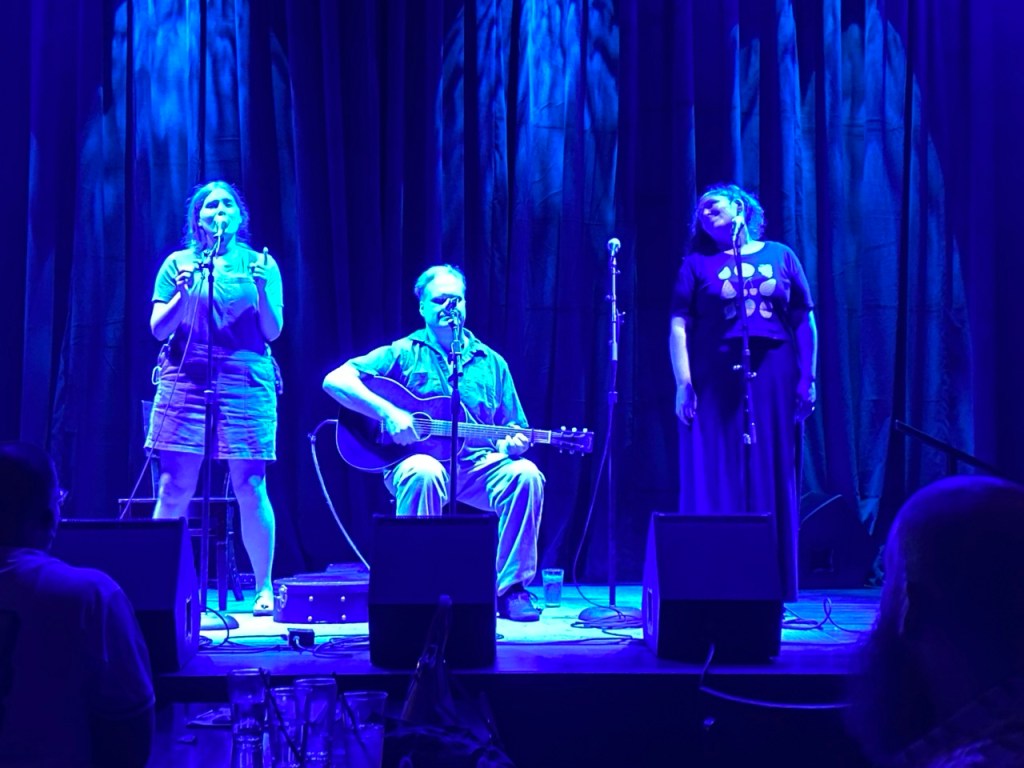 A photo of 3 people performing on a stage. Two people are singing, and one is playing guitar.